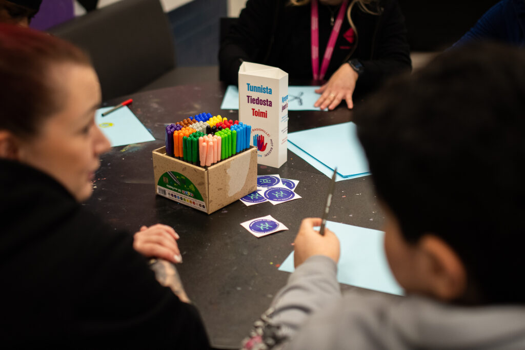 Nuori ja nuorisotyöntekijä pöydän ääressä. Pöydällä on kyniä ja osaamiskeskuksen tarroja.